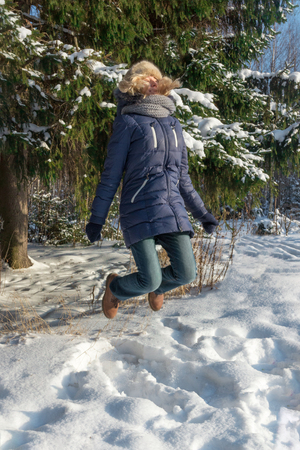 Happy young woman in fur hat jumps near green fir tree outdoor in sunny winter dayの写真素材