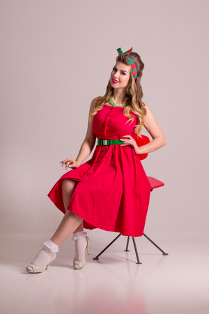 Happy girl in red dress with hairdo sits on chair in grey studio, pin up styleの写真素材