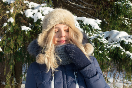 Pretty young woman in hat poses with closed eyes near green fir tree outdoor in sunny winter dayの写真素材