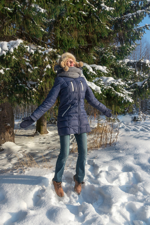 Happy girl in fur hat jumps near green fir tree outdoor in sunny winter dayの写真素材