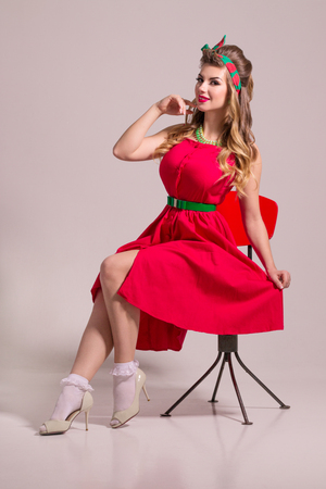Smiling girl in red dress with hairdo sits on chair in grey studio, pin up styleの写真素材