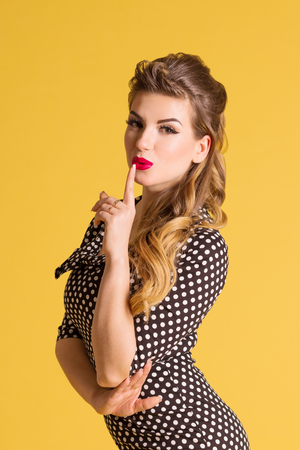 Beautiful happy woman shows quiet gesture in yellow studio, pin up styleの写真素材