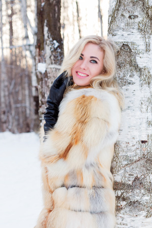 Blonde in fur coat, leather gloves smiles near birÑh outdoor at winter day in forestの写真素材