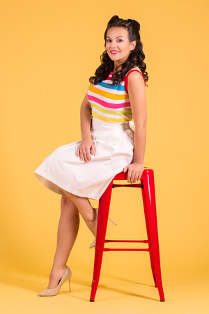 Smiling woman in skirt with hairdo poses on stool in yellow studio, pin up style, full bodyの写真素材
