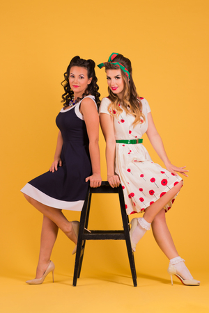 Two young women in dresses pose in yellow studio near stool, pin up style, full bodyの写真素材