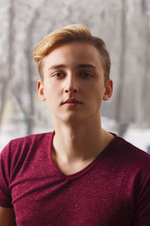 Handsome blond boy teenager in t-shirt poses  near window in roomの写真素材