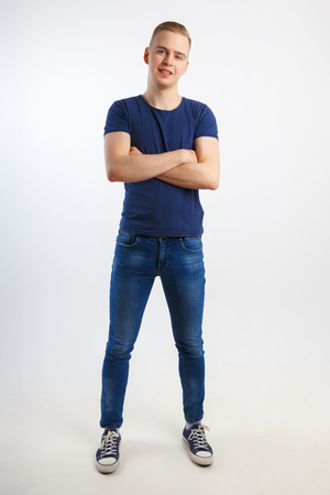 Young happy handsome man in blue shirt and jeans poses with crossed arms in white studio, full bodyの写真素材