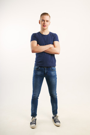 Young handsome man in blue shirt and jeans poses with crossed arms in white studio, full bodyの写真素材