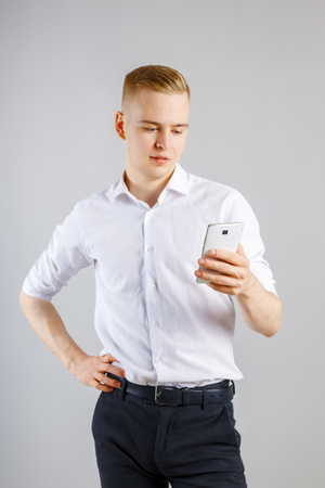 Young happy blond man in white shirt stands with smartphone in studioの写真素材