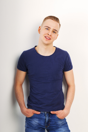 Young man in blue shirt and jeans smiles with hands in pockets in white studioの写真素材