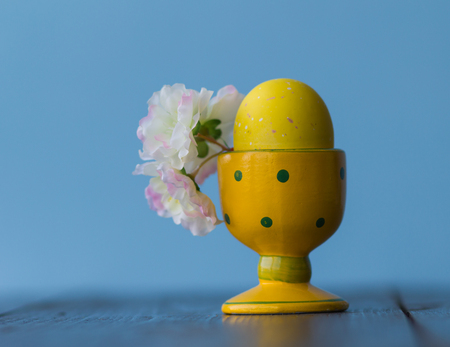 Closeup Easter yellow dotted egg cup with egg and flowers on the blue background.