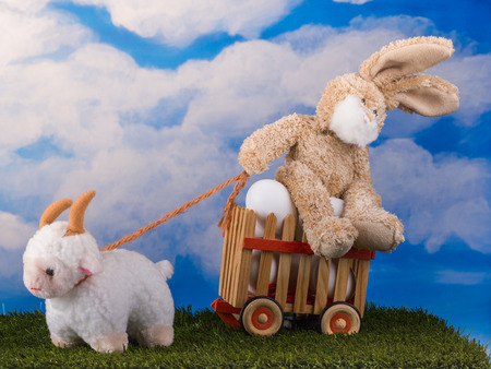 Funny toys bunny and lamb in scene with wooden cart of eggs on the blue sky background.の写真素材