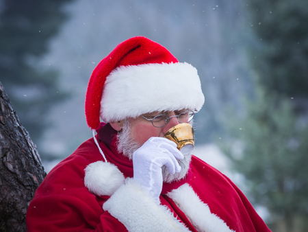 Portrait of traditional kindly Santa Claus with gold colored cup of coffee in the morning snow forest, close up.の写真素材