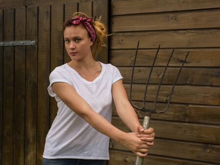 Charming young  attractive woman is standing by a wooden shed with a rake and looking at the cameraの写真素材