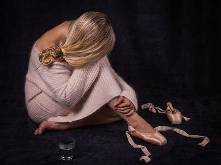 Wearied ballerina  with an inclined head, dressed in a warm pink sweater,  sits on the floor with loose ballet shoes, glass of water, on the black background.の写真素材