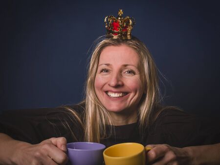 Smiling happy woman in crown keeps two cups of coffee in her hands, on the blue background.の写真素材