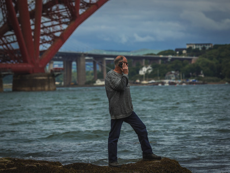 Senior modern man standing at river coast by  Edinburgh Bridge and talking on mobile.