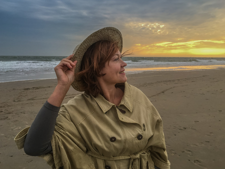 Portrait of smiling woman in straw hat and summer coat on the seacoast against the sunset skyの写真素材