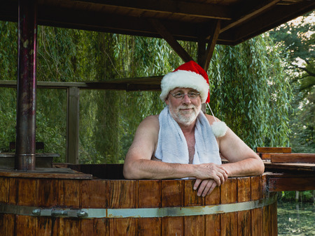 Senior man as Santa Claus with towel standing in a bath with warm water outside and looking with smiling at the camera.の写真素材