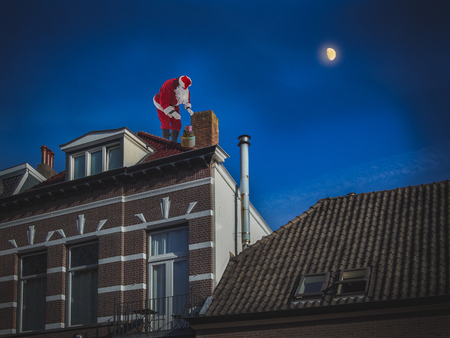 Santa Claus on the roof of a house puts the presents in the chimney. Christmas night scene, on the background of dark blue sky with a moon.の写真素材