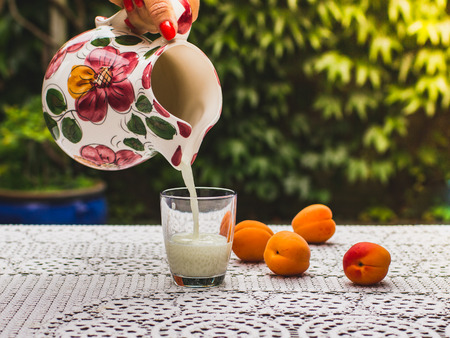 A woman pours milk from a ceramic jug with flower patterns into a drinking glass in the garden. Ripe peaches are lying on the table with a white tablecloth.の写真素材
