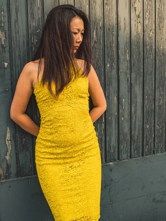 Pensive Asian model of 40 years in a yellow lace dress on the background of a rough textured wooden...