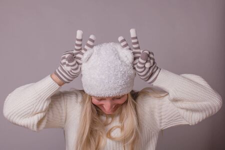 Humorous woman wearing season winter clothes, with long blond hair, makes horns on her head with fingers in knitted striped gloves, pastel colors over gray-lilac background.の写真素材