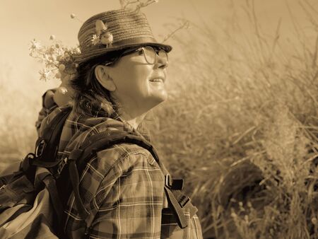 Mature woman in a hat and with a backpack surrounded by nature looks up.の写真素材