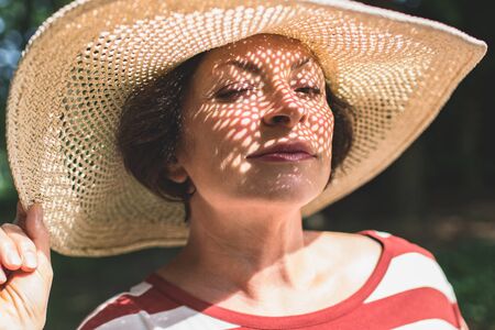 Closeup portrait of mature charming lady in big straw hat with winking eye. Summer vacation, sunny day, outdoorの写真素材