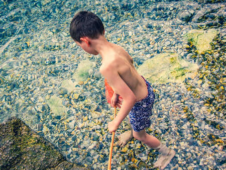 Cute little boy of 6 years playing with fishing net looking for crabs in sea water on a background of stones. Top view from back.の写真素材