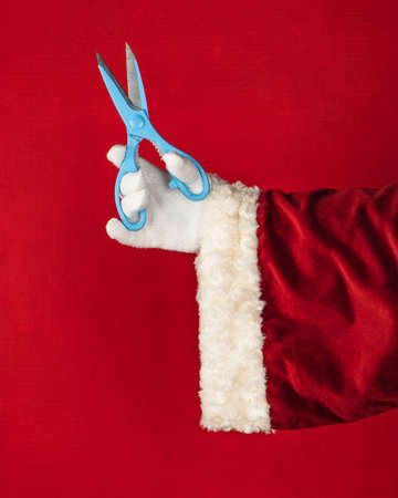 Close-up hand of Santa Claus holding in hands blue scissors isolated on bright vibrant red color background with copy space.の写真素材
