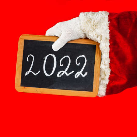 Santa Claus's hand is holding a blackboard with the new year 2022 number written in chalk. Red background and red Santa Claus's costume.の写真素材