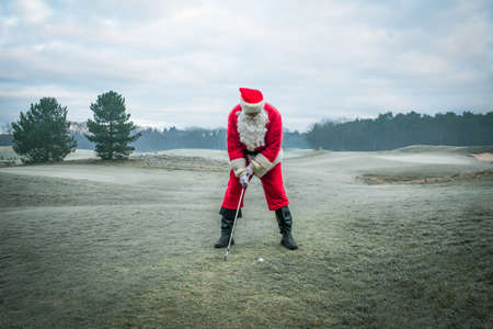 Santa Claus in a red traditional costume plays golf on a green frosty field, outside in winter.の写真素材