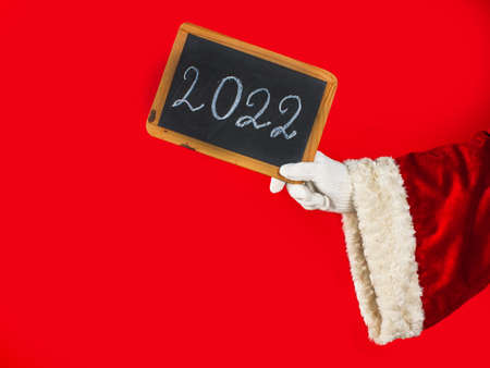 Close up of the hand of Santa Claus with a blackboard where the numbers of the new year 2022 are written in chalk. The image on a red background with space for text. New year concept.の写真素材