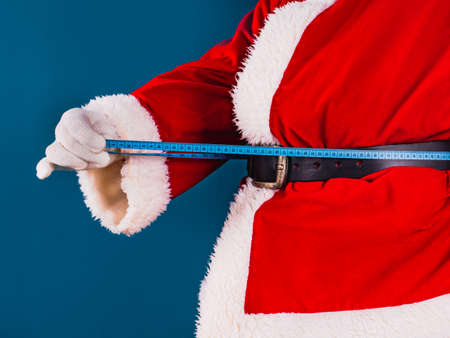 Fat Santa Claus stands on a blue background and holds a centimeter in his hand, tries to measure his waist.の写真素材