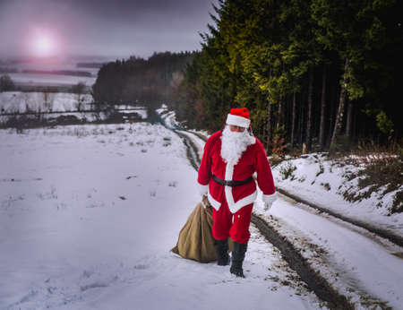 A man in the traditional costume of Santa Claus walks along the road, dragging a heavy jute bag with gifts. Long way, evening, tired.の写真素材