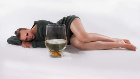 Young woman with long legs and in a green sweater lies on a white background in the studio and looks thoughtfully at a goldfish in an aquarium. Taking care of pets.の写真素材