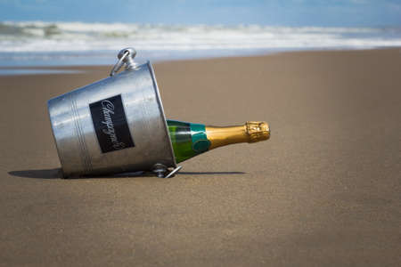 A closed bottle of champagne in a champagne bucket lie on the wet sand on the seashore.の写真素材