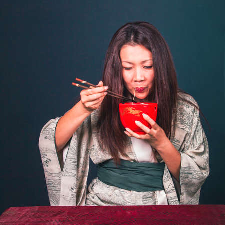 Beautiful cute Asian woman eating asian noodles with wood chopsticks. studio shot on a dark green backgroundの写真素材