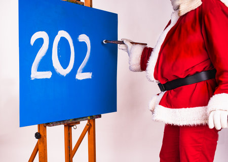 Santa Claus in the form of an artist writes the numbers 2023 or other numbers with white paint on a blue board that stands on an easel in the studio.の写真素材