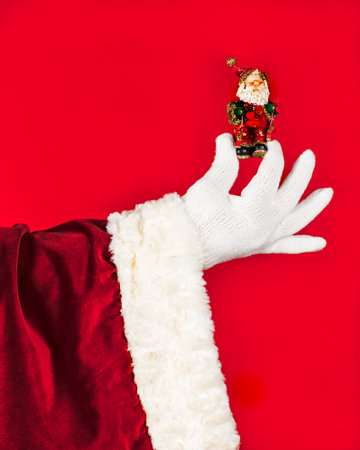 The hand of Santa Claus in a luxurious suit and a white glove holds in his hand a souvenir, a statuette of Santa Claus on a red background, with space for copy.の写真素材