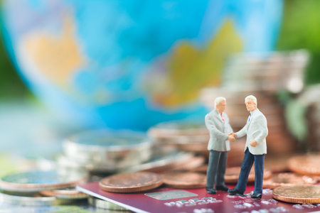 Miniature businessman handshaking and standing on the credit card or debit card with blurred globe and stacks of coins background using as commitment, agreement, investment, partnership , E-commerce and online business concept.の写真素材
