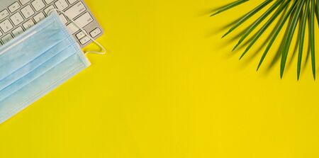 Top view of yellow color office table with surgical face mask, keyboard and palm leaves. Copy space.