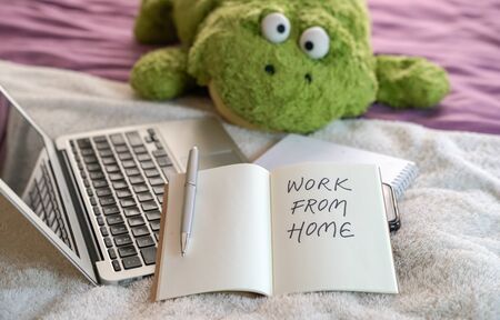 Work from home, text written on notepad. Computer laptop, soft toy and notepad on top of bed in the...