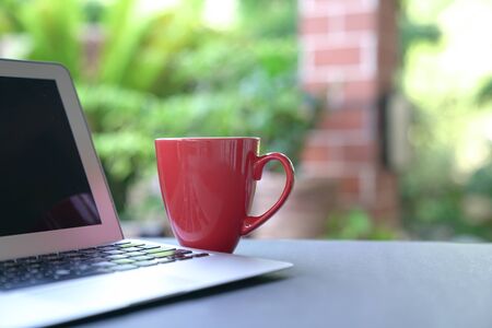 Computer laptop and coffee in red cup with garden background. Work from home. Copy space.の写真素材