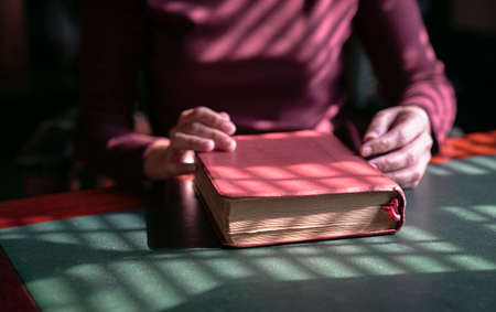 Woman's hands on a closed bible on a table. Artistic lines patterns of light and shadow.