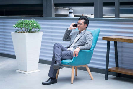 Chinese businessman sitting on a chair and having a phone call via his cellphoneの写真素材