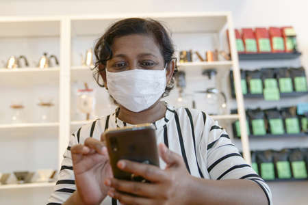 Indian woman with face covered with medical face mask reading her cellphone inside a restaurant.の写真素材