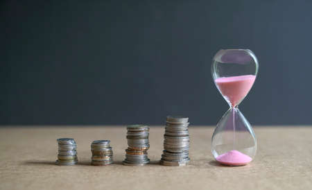 Hourglass with sand flowing and stacked coins on table. Time is money concept.