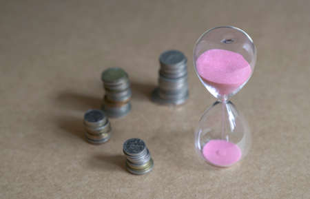 Hourglass with sand flowing and stacked coins on table. Time is money concept.